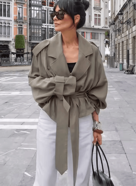 25 Minimalist Capsule Wardrobe Essentials 2026 | Timeless Pieces Every Woman Needs 15 A model wearing a sophisticated, French-style khaki windbreaker with a dramatic big collar and waist-tie detail, styled for a modern and chic 2026 autumn commuter look.
